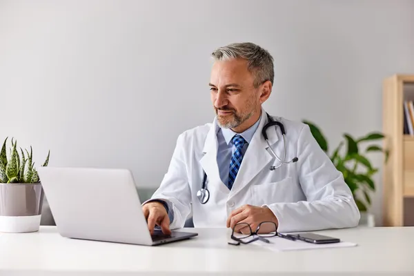 A smiling male doctor working over the laptop, sitting in the modern office