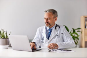 a smiling male doctor working over the laptop, sitting in the modern office.